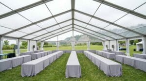 Interior of a large clear-span tent with rows of long banquet tables draped in grey linens and glass walls revealing a scenic landscape