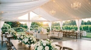 Stunning clear-span wedding tent interior with white draped fabric ceiling, crystal chandeliers, lounge seating, long harvest tables, and floor-to-ceiling vineyard views at sunset