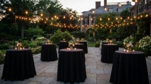 Dusk cocktail party on a stone patio at a garden estate with black tablecloths, floral centerpieces, and warm string lights overhead