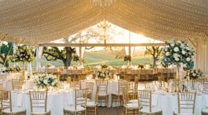 Sunlit marquee tent interior with white tablecloths, gold chiavari chairs, tall floral centerpieces, and fairy light ceiling