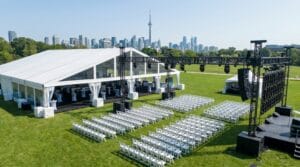 Aerial view of an outdoor corporate event with a white frame tent, professional stage, lighting rigs, and rows of white chairs near the Toronto skyline