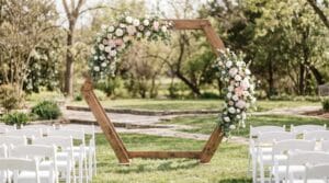 Hexagonal wooden ceremony arch adorned with blush pink and white roses flanked by rows of white folding chairs on a lawn