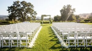 Sunlit outdoor wedding ceremony aisle with rows of white folding chairs, rose petal path, and a lush green floral arch at the altar with rolling hills
