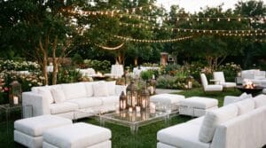 Outdoor event lounge with white sofas, ottomans, Moroccan lanterns, a fire pit, and bistro string lights canopying a lush garden at dusk