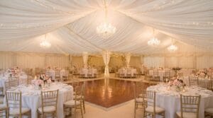 Luxurious marquee tent interior with white fabric draping, crystal chandeliers, fairy lights, gold chiavari chairs, and pink floral centerpieces