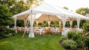 White pole tent in a lush backyard garden at sunset with round tables, folding wooden chairs, and colorful floral centerpieces