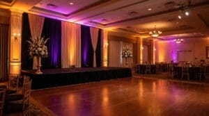 Grand ballroom with a raised stage, dark curtain backdrop, tall floral arrangements, polished dance floor, and amber and purple uplighting