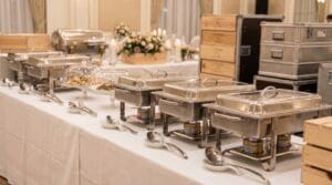 Professional catering buffet table with stainless steel chafing dishes, serving utensils, and elegant floral arrangements in a banquet hall