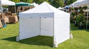 White pop-up canopy tent with sidewalls on grass at an outdoor festival or market with vendor stalls in the background