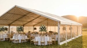 Wide-angle view of an outdoor wedding tent at golden hour with glass sidewalls, white tablecloths, gold chiavari chairs, tall florals, and fairy lights