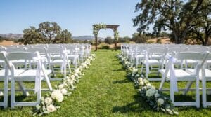 Wide-angle view of an outdoor wedding ceremony with rows of white chairs, a floral and greenery arch, and rolling hills in the background