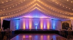 Elegant tent interior with a raised stage, white draping, and purple and blue uplighting over a spacious dance floor with gold chiavari chairs